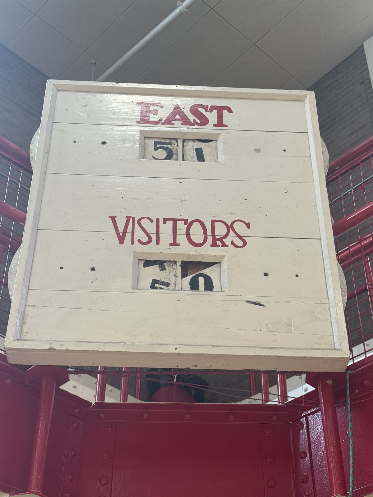 The original hand-operated wooden scoreboard still mounted in the East High School gymnasium, its cream-painted boards lettered EAST and VISITORS in red above sliding number panels, set against the gym's red steel framework